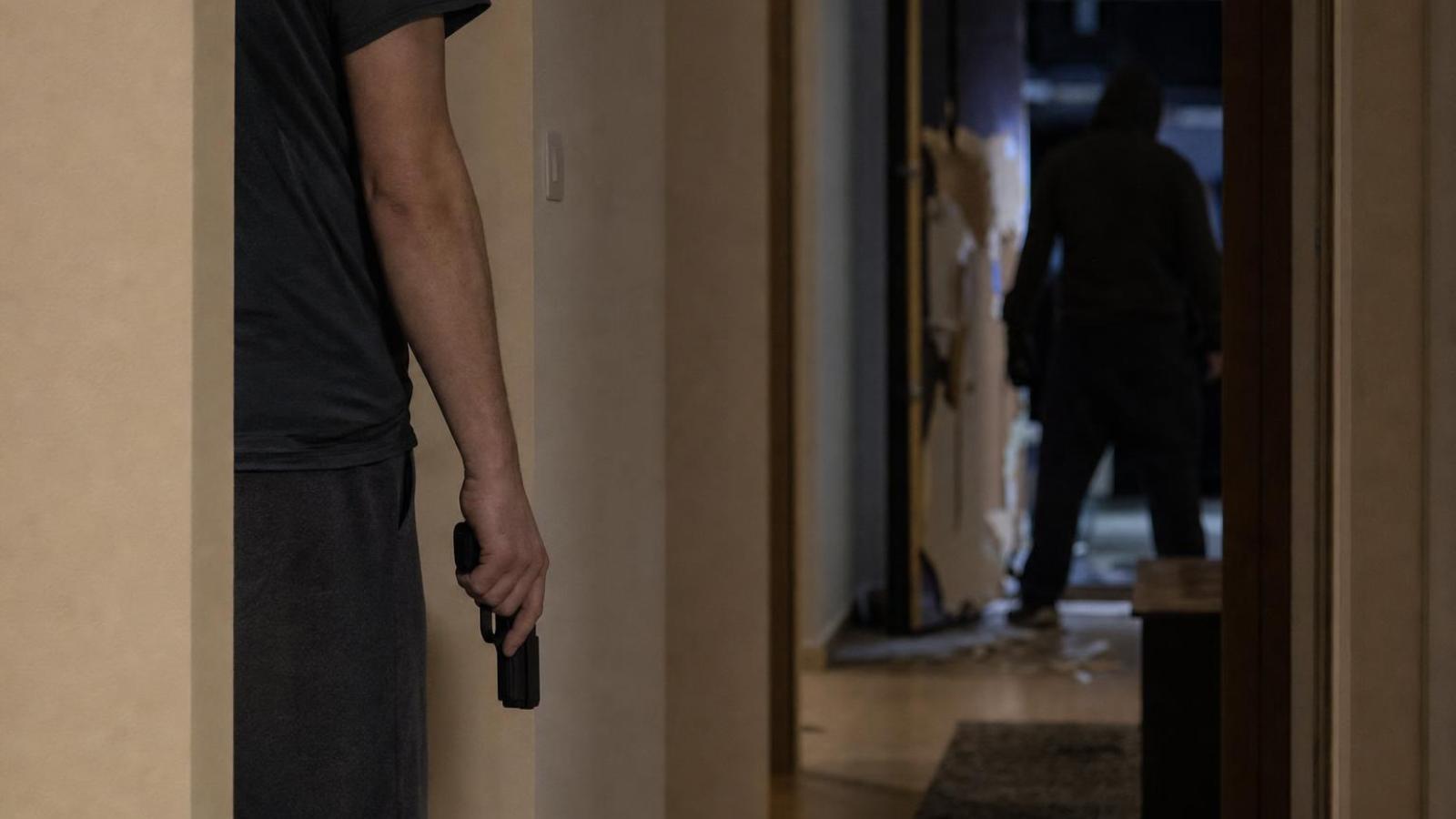 Ozbrojený obyvateľ v chodbe domu pri poškodených dverách a siluete votrelca.